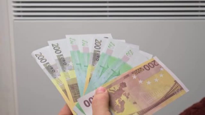 在家中电加热器散热器附近的人手拿着欧元钞票。男人付钱取暖。加热昂贵。节能。生活成本上升。全球天然气危