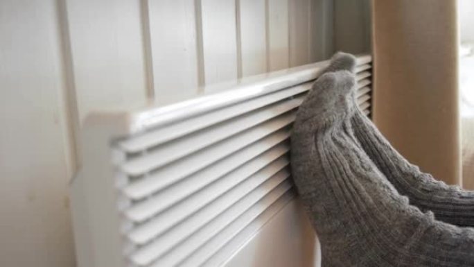 针织袜子里的腿在电加热器上变暖。冻着的女人穿着温暖的羊毛袜冻着冬天的寒冷。呆在家里不舒服。女孩在现代