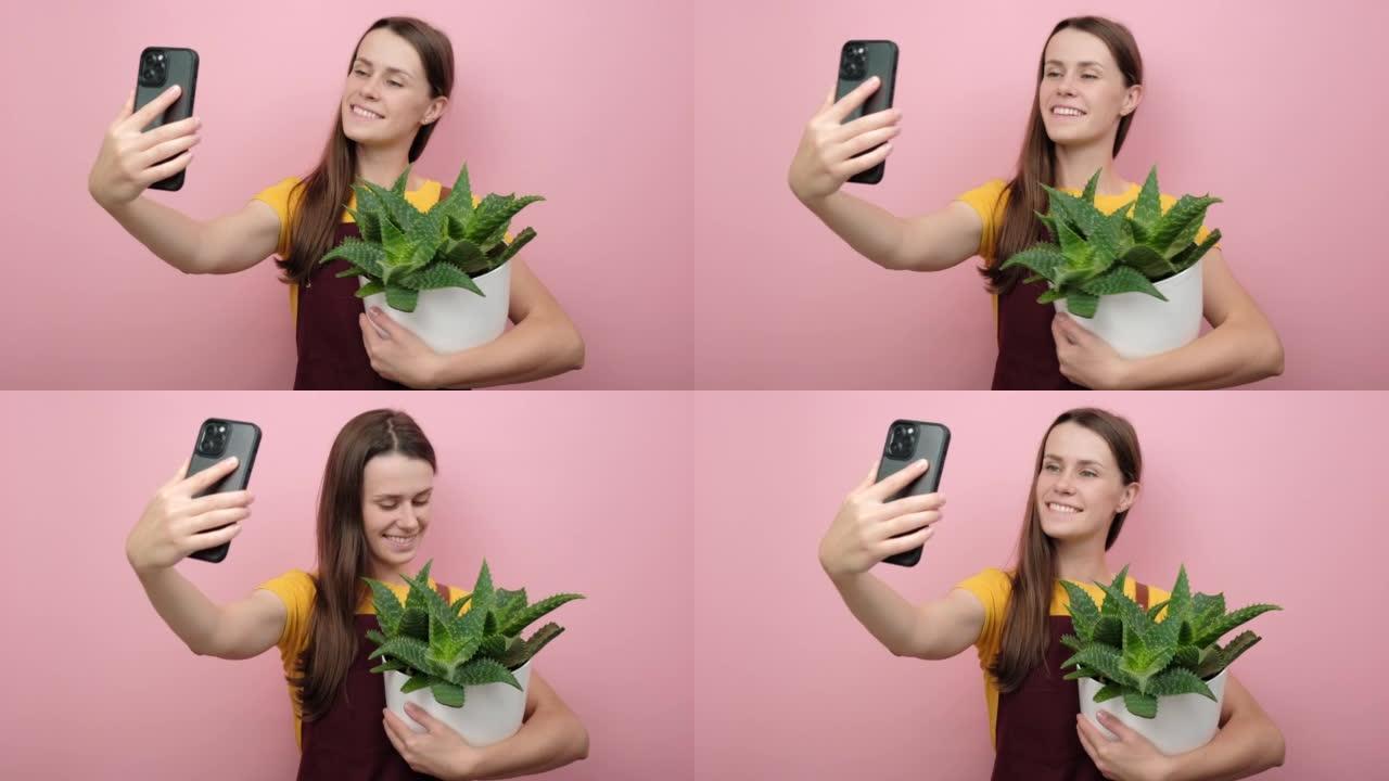 微笑的年轻女子花店博客作者在智能手机相机上与花盆中的植物合影，孤立在粉红色背景上。园艺，家庭主妇，在