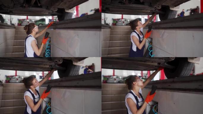 女机械师用遥控器检查液压车轮播放检测器上的汽车悬架。坚强的女人检查行走装置和转向部件的磨损。穿着工作