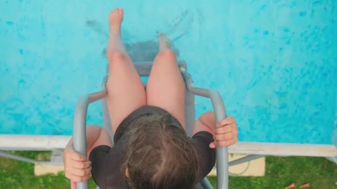 穿着泳衣的快乐高加索女孩坐在花园泳池梯子上，用脚泼水