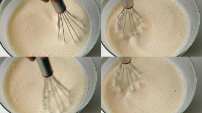 在厨房里用搅拌器在玻璃碗中搅拌煎饼面糊的人。特写。慢动作。用手揉捏，搅拌面团。烹饪面团，顶视图。液体