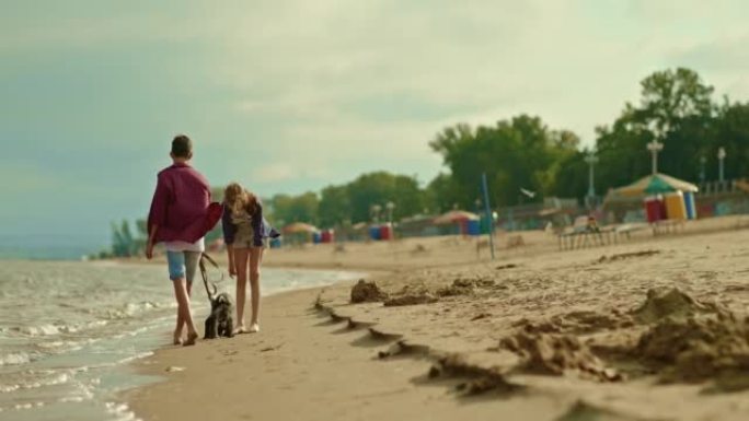 青少年带着狗沿着海滩散步，一个男人牵着狗。女孩兴高采烈地微笑着，走在河岸边。男孩和女孩赤脚走在潮湿的