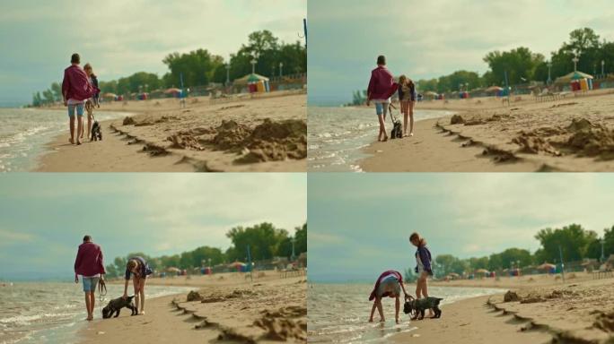 青少年带着狗沿着海滩散步，一个男人牵着狗。女孩兴高采烈地微笑着，走在河岸边。男孩和女孩赤脚走在潮湿的