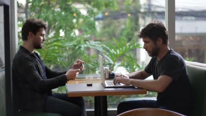 坐在咖啡店里使用技术的人。男性企业家在笔记本电脑上打字。同事使用智能手机浏览互联网在cafe pla