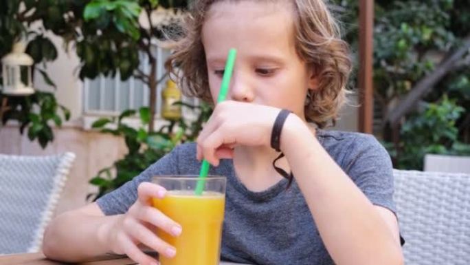 可爱的卷发男孩在户外咖啡馆的餐桌上喝鲜榨柑橘汁。小男孩喜欢橙汁。