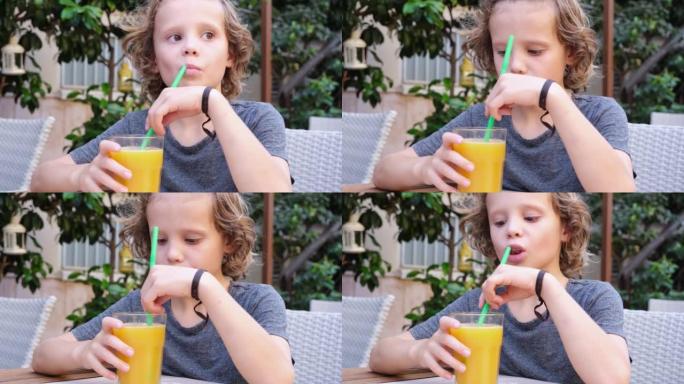 可爱的卷发男孩在户外咖啡馆的餐桌上喝鲜榨柑橘汁。小男孩喜欢橙汁。