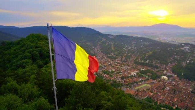 国旗位于山顶上，绿树成荫。罗马尼亚日落时背景上有多座建筑的布拉索夫视图