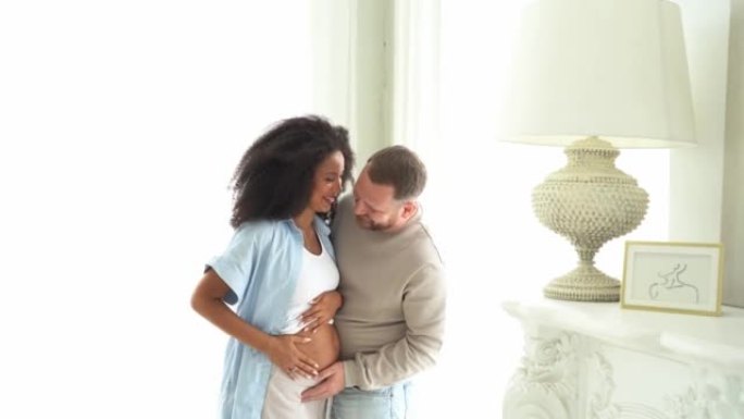 白人摸着他怀孕的黑人妻子的肚子。孕妇和慈爱的丈夫在家里拥抱肚子。多民族夫妇在怀孕的腹部感受婴儿的运动
