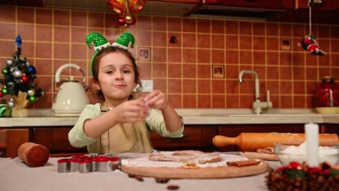 带着精灵箍和厨师围裙的漂亮女孩正在从姜饼面团上切割模具，并向相机展示