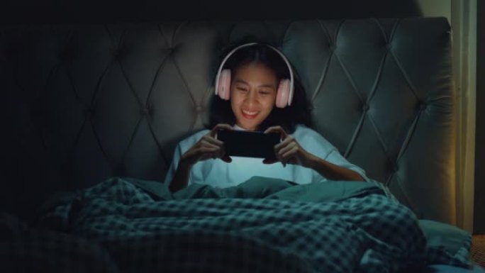 年轻的亚洲女孩媒体成瘾在床上戴着耳机笑在流媒体在线视频点播中观看最喜欢的电影，晚上在家点播视频。