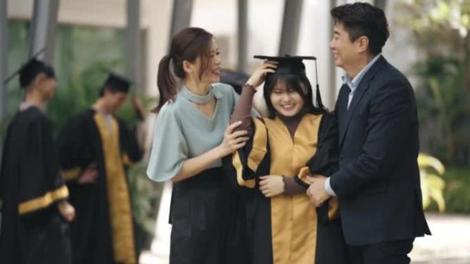 自豪的亚洲华裔父母在大学大楼外的大学毕业礼服中调整女儿的迫击炮