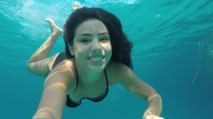 穿着比基尼的美丽年轻女子在水下游泳，对着可穿戴相机微笑，从海面上走出来