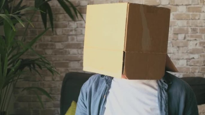 成熟的男人穿着空纸箱，坐在房子客厅的沙发上。高加索人，脸藏在纸板送货容器里，在家沙发上放松