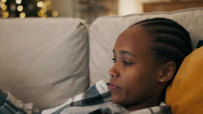 一个心烦意乱的非洲女孩穿着格子衬衫，躺在客厅的沙发上看电影。她感动，嘴唇和对电影英雄的担忧