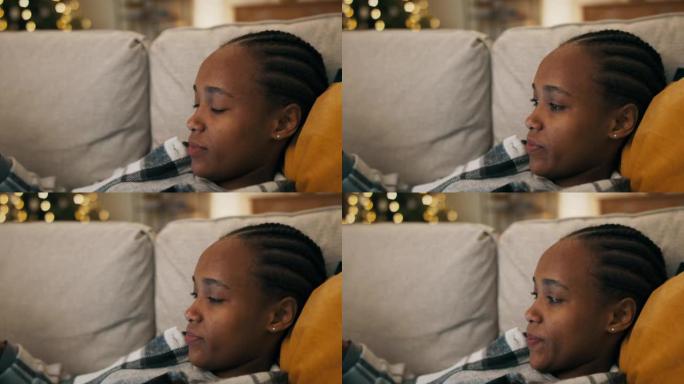 一个心烦意乱的非洲女孩穿着格子衬衫，躺在客厅的沙发上看电影。她感动，嘴唇和对电影英雄的担忧