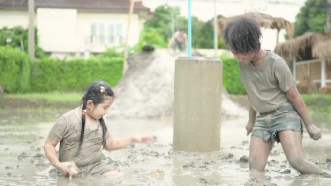 孩子们在社区田野里的泥地里玩耍，在泥泞的田野里抓青蛙，玩得很开心。父母从远处看着