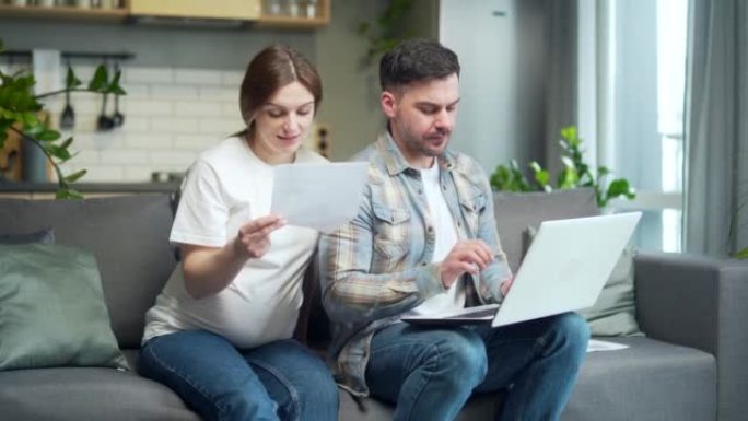 年轻的已婚夫妇，丈夫和怀孕的妻子一起在家里，满意地坐在沙发上，在笔记本电脑上工作，在家里使用网站在线