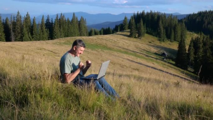 一个男人坐在山上美丽的草地上，在笔记本电脑上工作