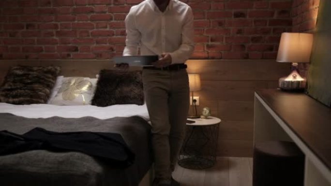 穿着正式白衬衫的英俊年轻商人坐在现代房间里的平板电脑床上。独资经营者躺在酒店的床上，在平板电脑上工作