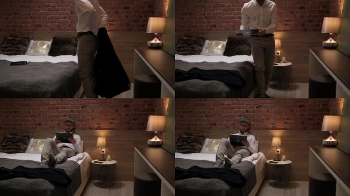 穿着正式白衬衫的英俊年轻商人坐在现代房间里的平板电脑床上。独资经营者躺在酒店的床上，在平板电脑上工作