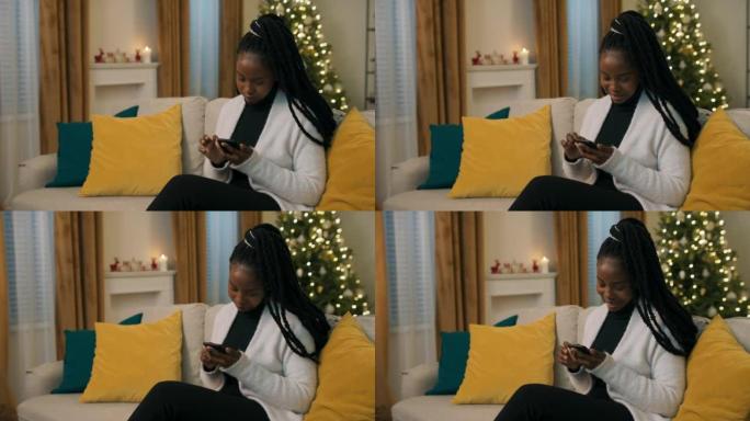 冬天的晚上。一个非洲外表漂亮的女孩手里拿着手机，为家庭相册选择照片。女孩穿着黑色衣服和白色开衫。沙发