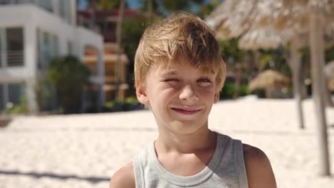 海滩上一个年轻的白人金发男孩的肖像。他眯着眼睛看着太阳，看着相机