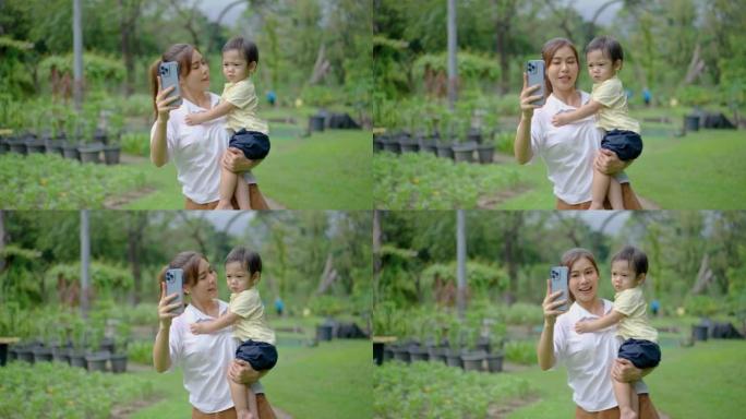 公园的家庭娱乐和周末休闲。亚洲妈妈和她的小儿子在绿色夏日公园自拍，妈妈用手机拍照，他们高兴地对着镜头