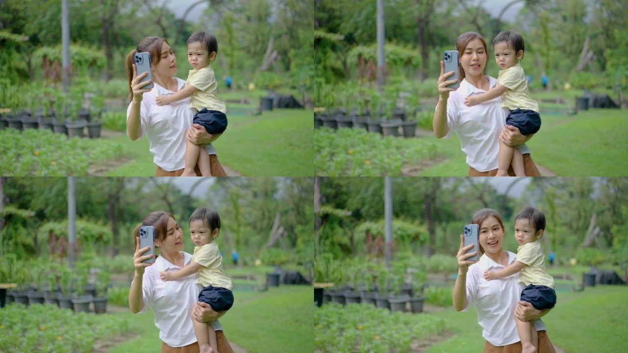 公园的家庭娱乐和周末休闲。亚洲妈妈和她的小儿子在绿色夏日公园自拍，妈妈用手机拍照，他们高兴地对着镜头