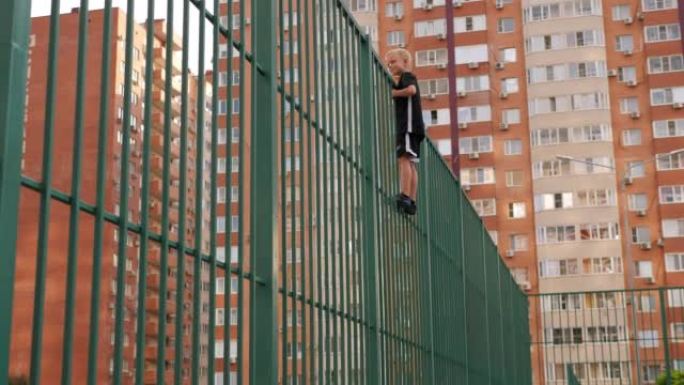 一个男孩在建筑物背景下的运动场上爬上高高的篱笆。