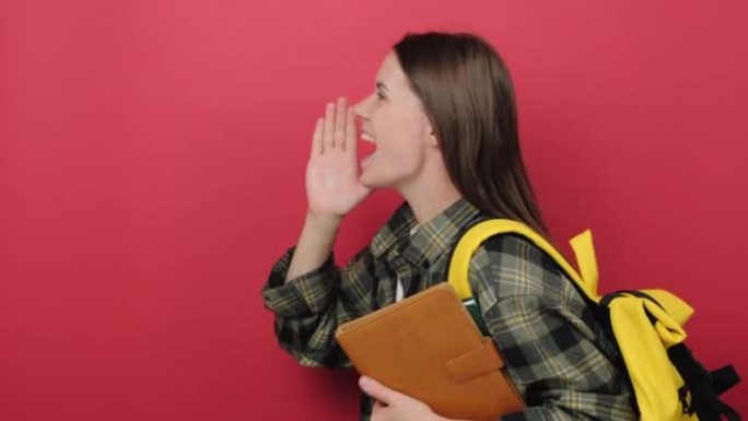 20多岁的年轻学生女子肖像，黄色背包拿着两本书向侧面喊着，宣布着什么，在工作室的红色背景墙上孤立地摆