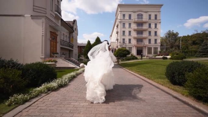 漂亮的新娘穿着长长的通风面纱，优雅的白色连衣裙在婚礼当天远离相机。顽皮迷人的女人逃跑转身，看着观众。