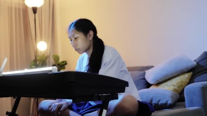 亚洲女孩在家里的夜晚通过沙发上的移动应用学习弹奏键盘乐器，生活方式理念。