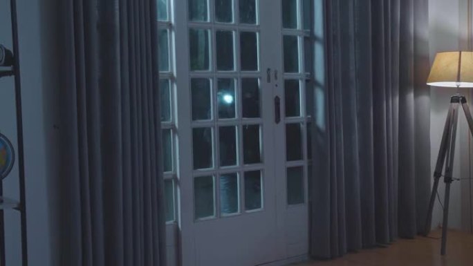 小偷男子拿着手电筒在漆黑的夜晚站在外面看着房子里