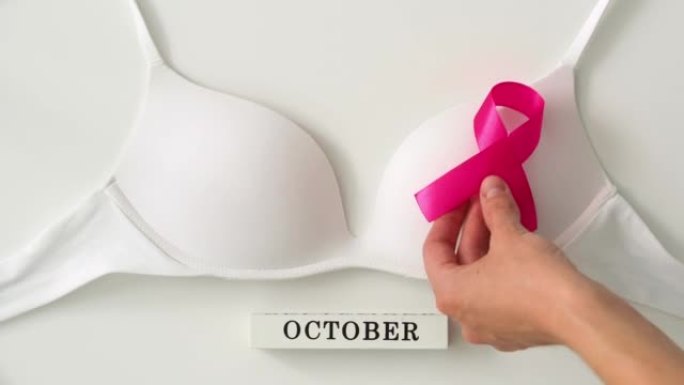 白色背景乳腺癌宣传月。女人的手放一条粉红色的丝带-白色胸罩上的蝴蝶结。希望和恢复的象征。世界癌症日概