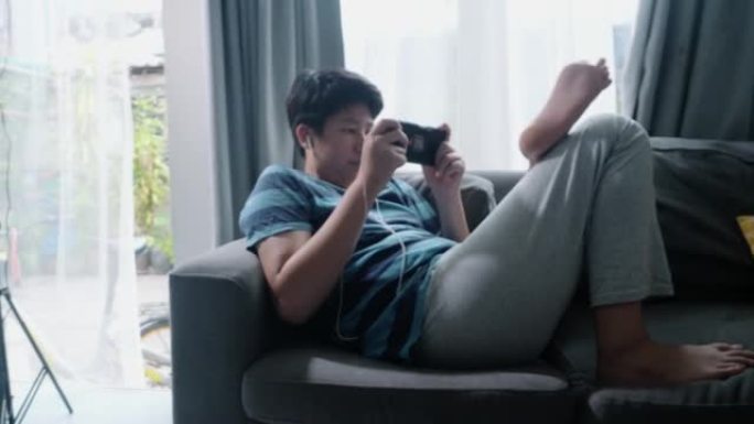亚洲少年男孩躺在沙发上，在家窗口附近玩手机游戏，生活方式概念。