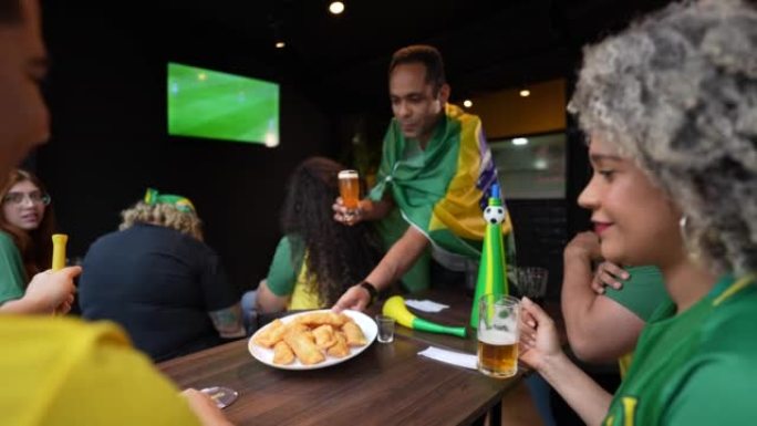 一名男子带着一盘粉彩和一杯啤酒给他的朋友在餐桌上看足球比赛