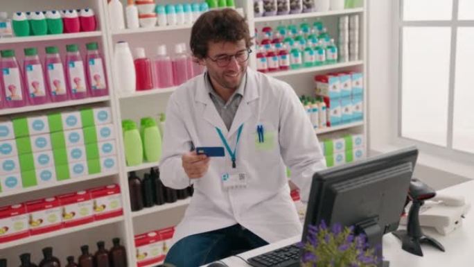 年轻的西班牙裔男子药剂师在药房使用持有信用卡的计算机