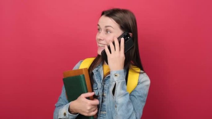 穿着牛仔夹克和黄色背包的快乐年轻女子学生拿着两本书在手机上说话，在红色背景墙上孤立地摆姿势。高中大学