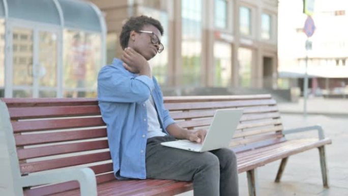 坐在户外长凳上使用笔记本电脑时颈部疼痛的非洲年轻人