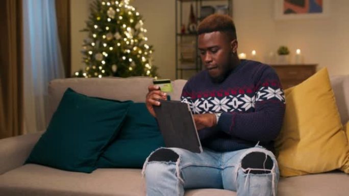 一个有吸引力的非洲人坐在沙发上，拿着平板电脑和银行卡，并为朋友订购圣诞节礼物。他输入卡片的详细信息。