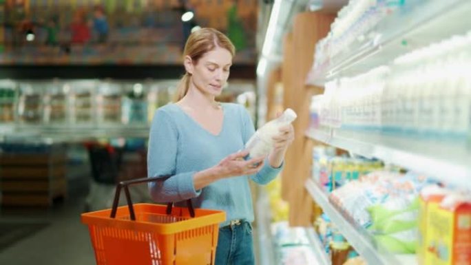 一名妇女选择乳制品，手里拿着一瓶牛奶。一位女士站在杂货店里拿着购物篮挑东西。研究并阅读标签上的信息。