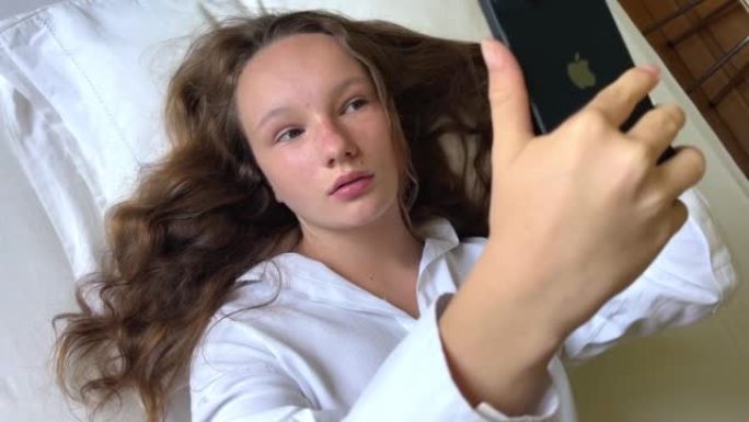 一个穿着白衬衫的女孩一个少年躺在白色的床上，手里拿着黑色的iPhone 13她看着屏幕平静地举起双手