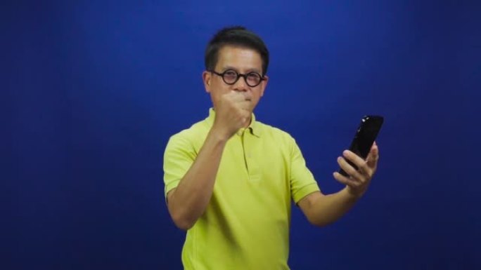 穿着休闲的黄色t恤的聪明的中年亚洲男人的肖像拿着智能手机，在工作室的蓝色背景上给迷你心孤立，看起来幸