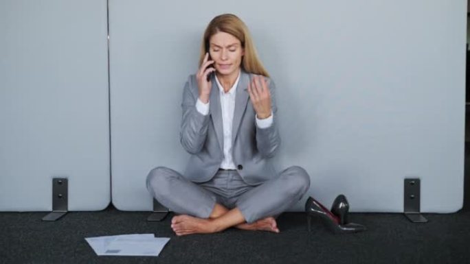 紧张疲倦的女商人坐在地板上脱鞋时正在打电话。工作中的问题。震惊沮丧的女商人学生感到压力很大，担心有不