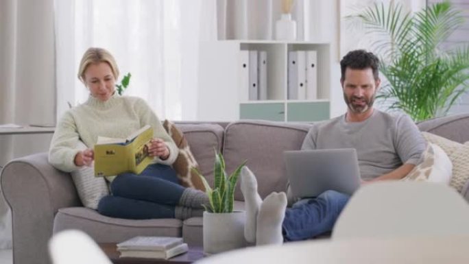 已婚现代中年夫妇一起坐在家里的客厅沙发上享受平静放松的生活方式。快乐，微笑的妻子看书，而丈夫则在笔记