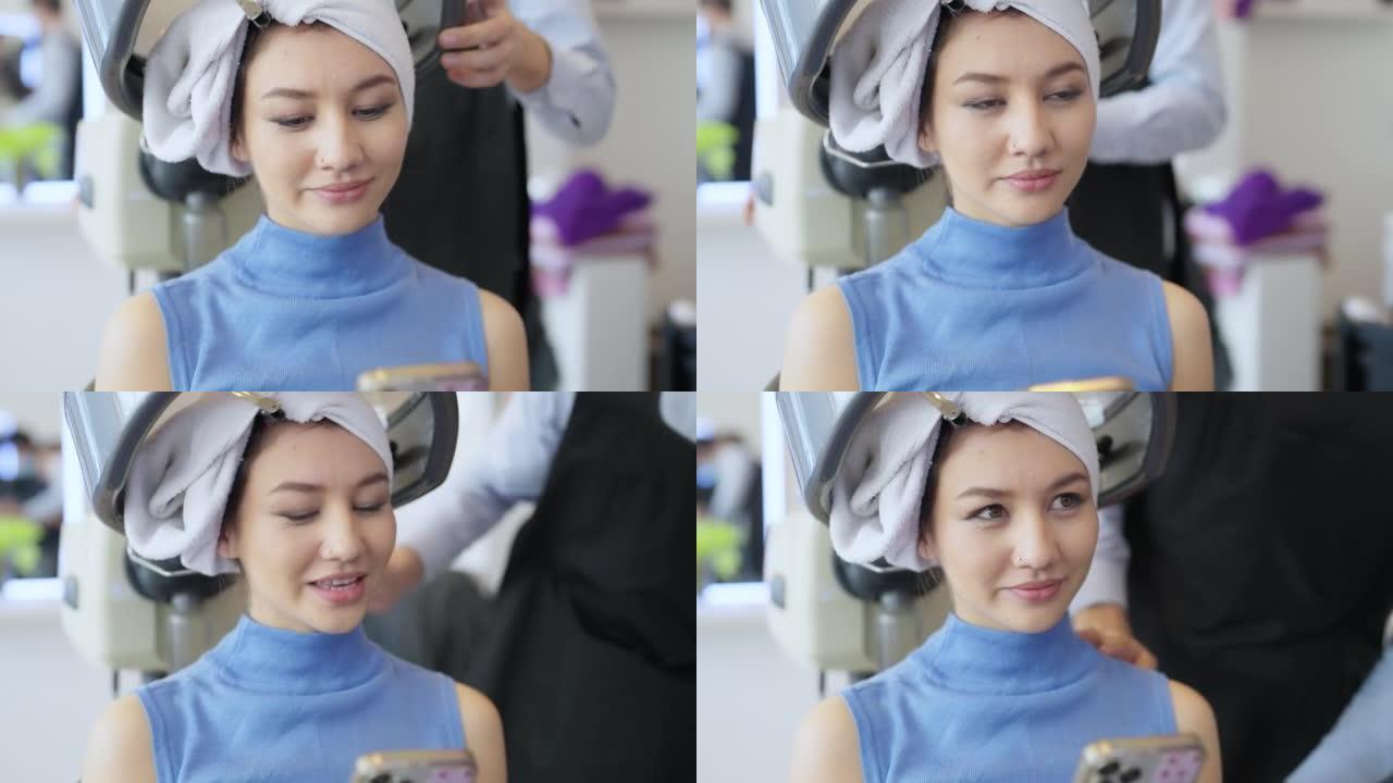 年轻女子的前视图在连帽烘干机下，手里拿着手机，在美容店染发。