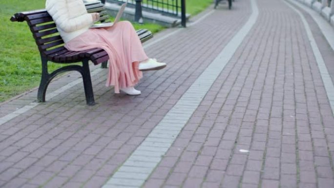 穿着白色外套粉色裙子的无法辨认的女性坐在长凳上使用笔记本电脑通话网络摄像头