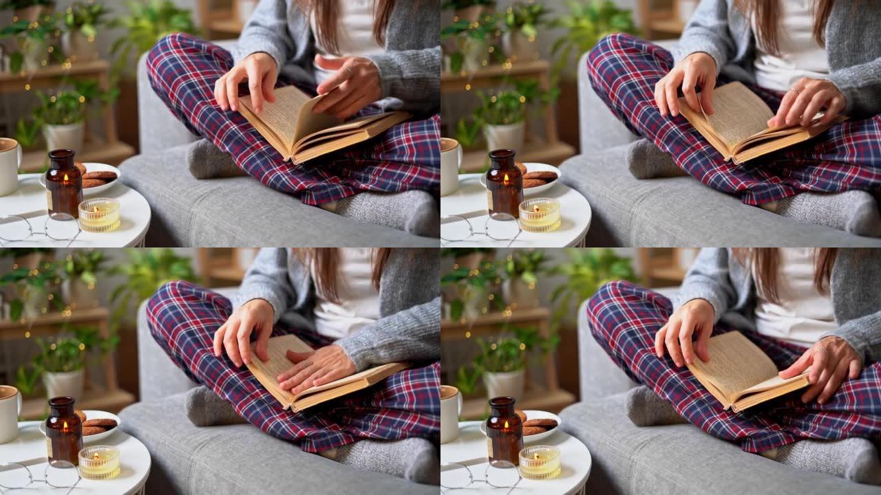舒适的女人穿着针织冬季保暖袜子和方格睡衣，看书，翻阅书本，在杯子里喝咖啡，坐在家里的沙发上。带有蜡烛
