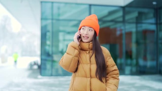 年轻的亚洲妇女在下雪的冬日里用手机说话，校园里的一名学生。快乐女孩对话站在城市街道上的现代城市背景